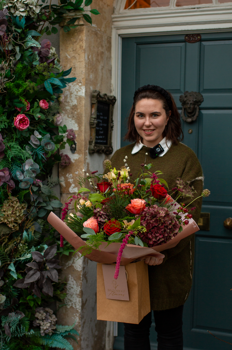 In front of a blue door, a person with floral design skills holds a vibrant bouquet, wearing a green sweater over a white collared shirt. Foliage and blooms beside them hint at their recent experience in Event's Handtied Bouquet Flower Workshop.