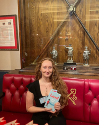 A woman in a black dress sits on a red cushioned bench, smiling with "In Conversation: Megan Clawson | Book Reading and Q&A" (Event) in hand. Behind her is a display case with swords, statues, and historical objects.