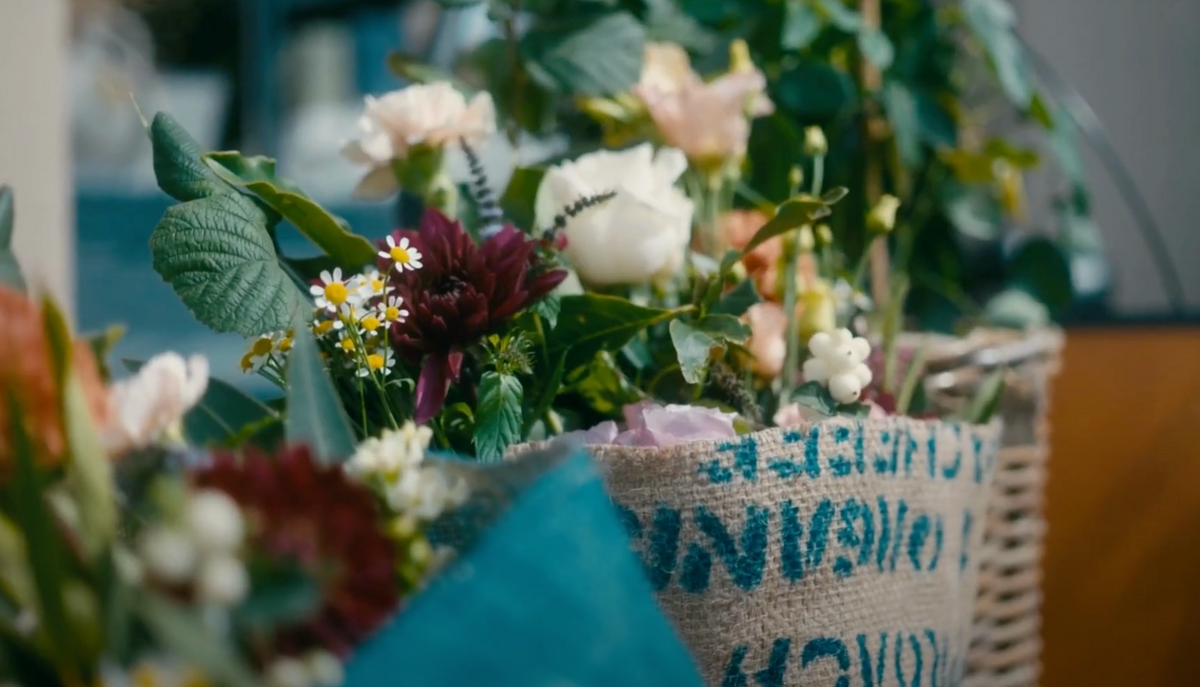A close-up of a woven basket filled with assorted fresh flowers and greenery, wrapped in burlap with blue text and teal fabric—a rustic, colorful display from Wild About Flowers' Friday Flowers Subscription.