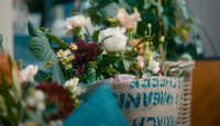 A close-up of a woven basket filled with assorted fresh flowers and greenery, wrapped in burlap with blue text and teal fabric—a rustic, colorful display from Wild About Flowers' Friday Flowers Subscription.