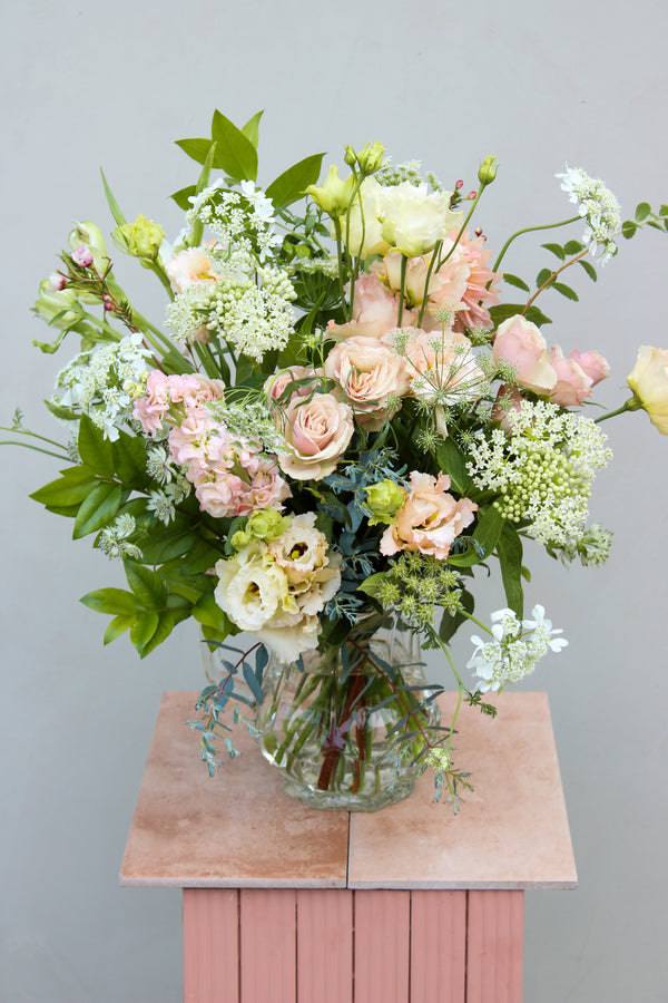 A clear glass vase, known as the Vintage Jug by Wild About Flowers, holds a flower arrangement of pastel blooms, featuring roses and lisianthus complemented by green foliage and delicate white flowers. This eco-conscious gift rests on a wooden stool against a light gray background, offering timeless elegance.
