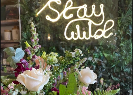 The Wild Wedding Bundle by Custom Bundle is a masterpiece with white roses, pink and purple flowers amidst lush greenery. A "Stay Wild" neon sign glows on a plant-covered wall, enhancing the captivating ambiance of this dummy product arrangement.