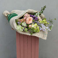 A bouquet artfully arranged in a Coffee wrap by Wild About Flowers, featuring pink roses, lavender, white wildflowers, and lush greenery. It is presented against a pale gray background on a ribbed muted pink pedestal.