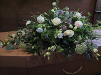 A 'From the Garden' Woodland Spray by Wild About Flowers decorates a funeral casket, featuring white roses, garden foliages, and delicate pastel flowers. The wooden casket is partially visible beneath the serene and respectful floral arrangement.