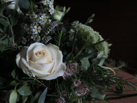 A close-up of the 'From the Garden' Woodland Spray by Wild About Flowers highlights a prominent white rose encircled by delicate white flowers, deep green leaves, and lush garden foliages, all set against a dark background.