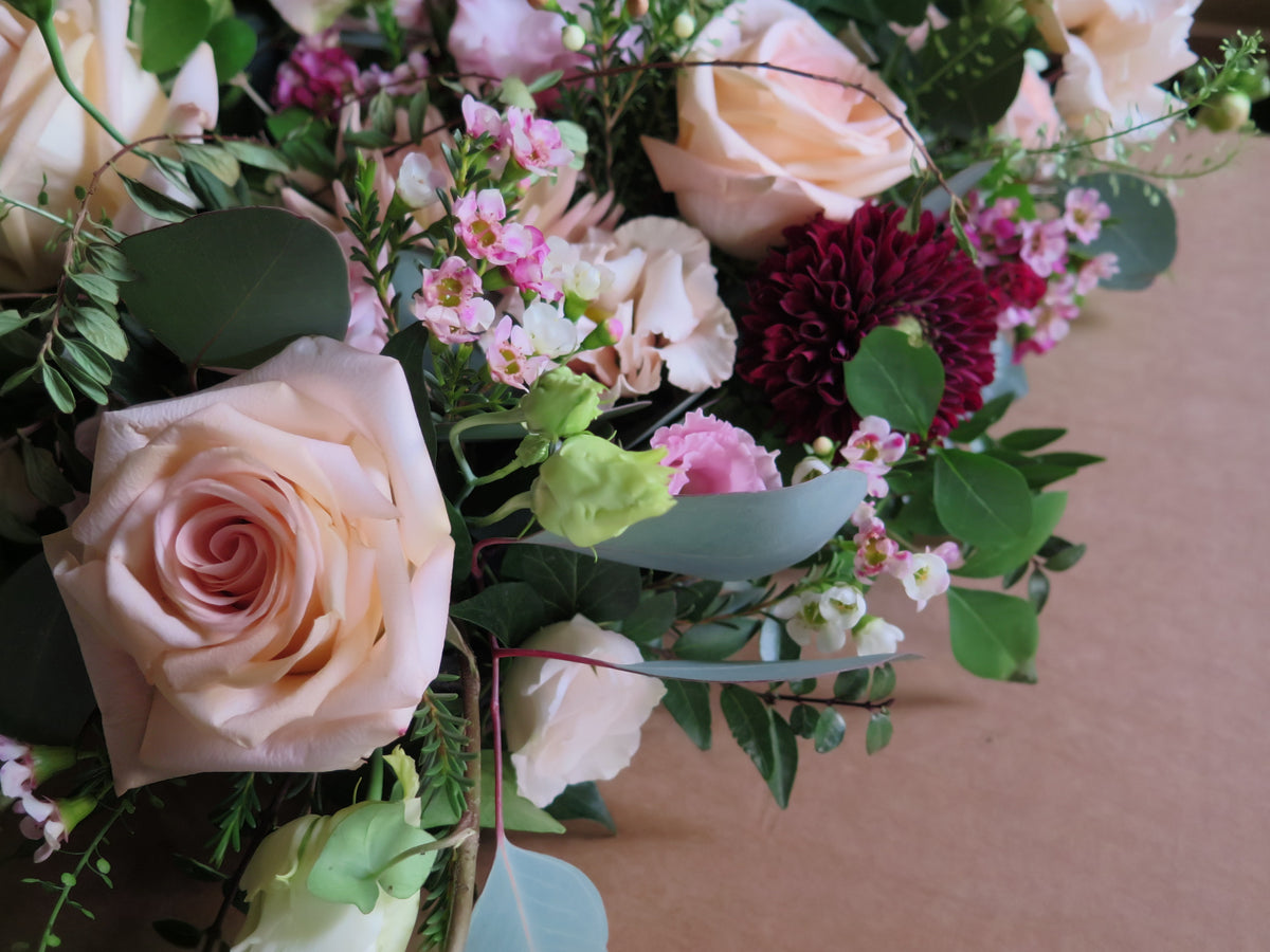 A detailed view of the 'From the Heart' Heart Design by Wild About Flowers showcases a floral arrangement with blush roses, deep red chrysanthemums, and delicate pink and white blossoms, all nestled amidst lush green foliage.