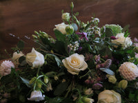 The 'From the Garden' Woodland Spray by Wild About Flowers showcases a lush combination of white roses, pink dahlias, and garden foliages set against a blurred brown background, creating a natural and elegant floral arrangement.