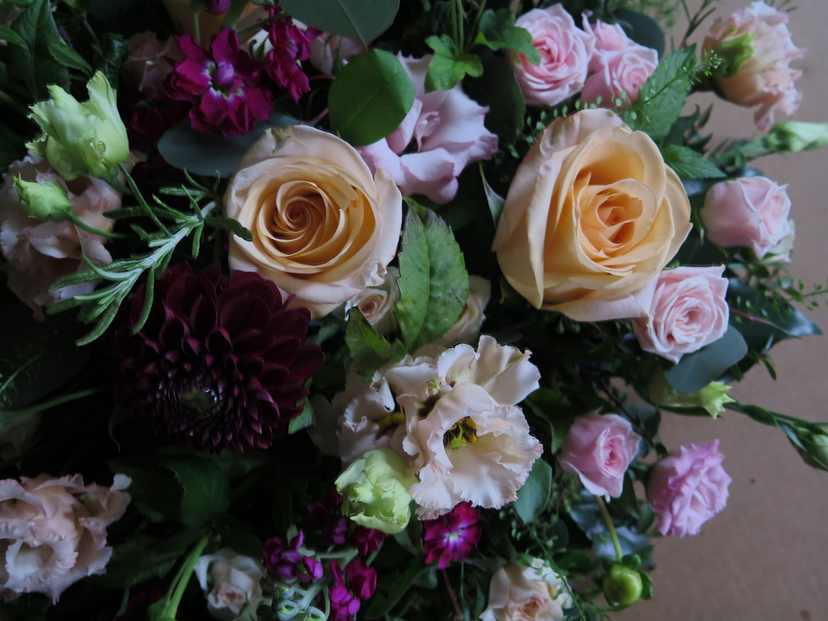 The 'From the Heart' Posy Design by Wild About Flowers showcases a delightful combination of pale yellow roses, pink roses, and an assortment of blooms in pink and purple hues, all enhanced with fresh cut foliage for a natural and elegant appearance.