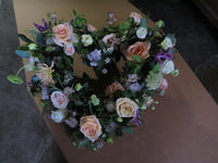 The 'From the Garden' Heart Design by Wild About Flowers features an elegant floral arrangement of lush pink and white seasonal roses, complemented by purple blooms and green foliage, beautifully presented on a brown surface.