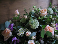 The 'From the Garden' Casket Spray by Wild About Flowers showcases a lush arrangement of pale pink roses, white flowers, and delicate purple blooms amidst green foliage. The soft, neutral brown background highlights the natural beauty of this tribute's seasonal flowers.