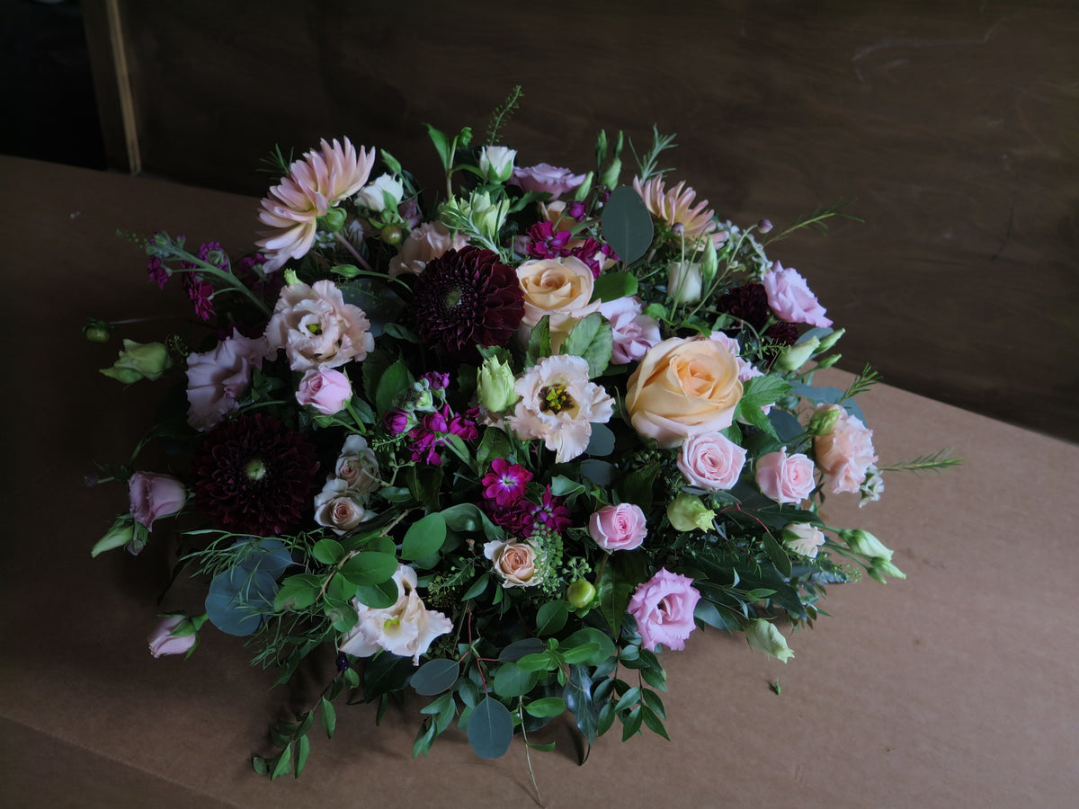 The 'From the Heart' Posy Design by Wild About Flowers is a funeral floral arrangement showcasing seasonal roses, dahlias, and lisianthus in pink, peach, and deep burgundy hues. It is enhanced with fresh cut foliage and presented on a simple brown background.