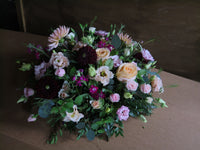 The 'From the Heart' Posy Design by Wild About Flowers is a funeral floral arrangement showcasing seasonal roses, dahlias, and lisianthus in pink, peach, and deep burgundy hues. It is enhanced with fresh cut foliage and presented on a simple brown background.