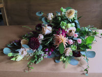 A luxurious and sustainable "From the Heart" Tied Sheaf by Wild About Flowers, featuring a vibrant assortment of roses, dahlias, and carnations in shades of pink, burgundy, and peach with lush green foliage displayed on a rustic brown surface.