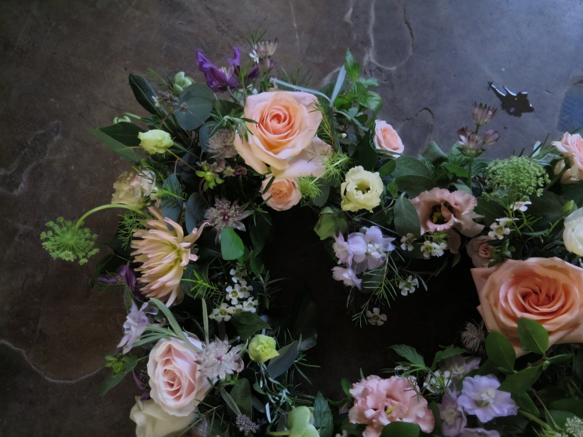 The 'From the Garden' Heart Design by Wild About Flowers showcases a beautiful arrangement of seasonal pale pink roses coupled with white and purple blooms, all elegantly set against a dark gray surface.