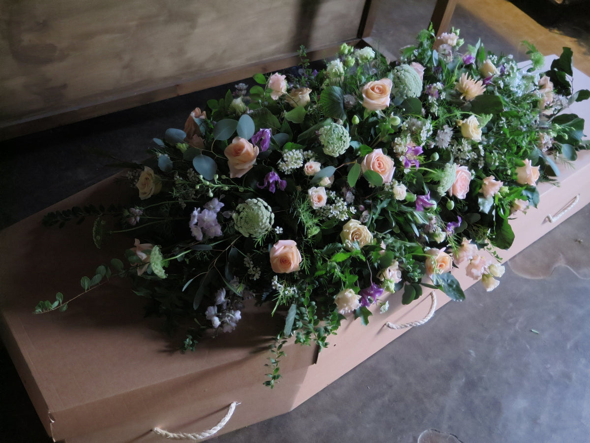 A cardboard coffin is embellished with the 'From the Garden' Casket Spray by Wild About Flowers, featuring a lush array of pink, white, and purple seasonal flowers including roses and greenery. It rests on a polished concrete floor, where soft, natural lighting highlights the tribute's delicate floral details beautifully.