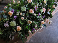 The 'From the Garden' Casket Spray by Wild About Flowers features a lush arrangement of pastel roses, seasonal flowers in purple and white, along with abundant green foliage. This serves as a heartfelt tribute amidst an ambiance enriched with soft lighting that conveys calm and respect.
