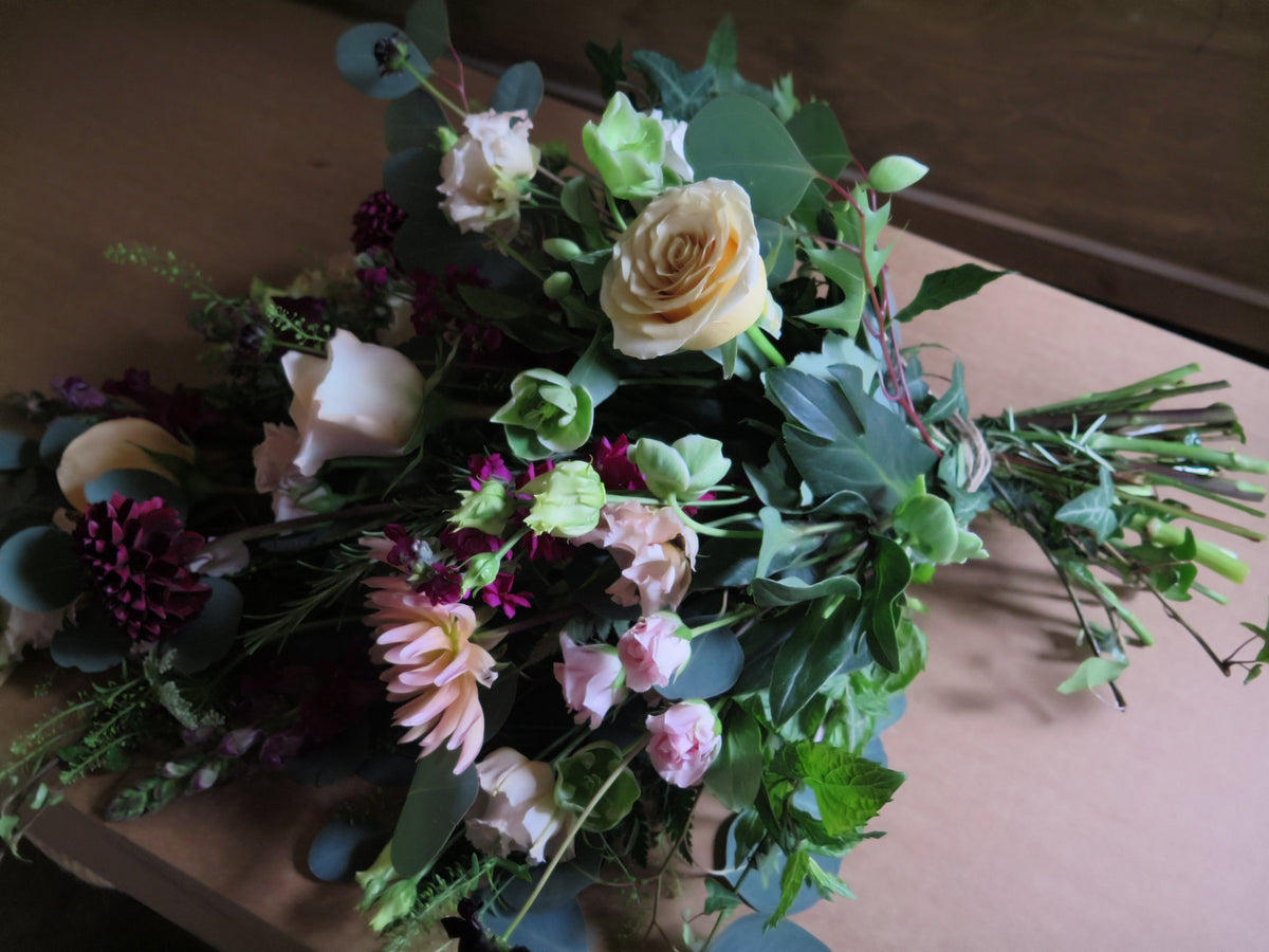 The 'From the Heart' Tied Sheaf from Wild About Flowers is an eco-friendly rustic bouquet featuring an assortment of roses, dahlias, and greenery. This stunning arrangement showcases a natural look with its blend of cream, pink, and dark purple tones set against a variety of greens on a wooden surface.