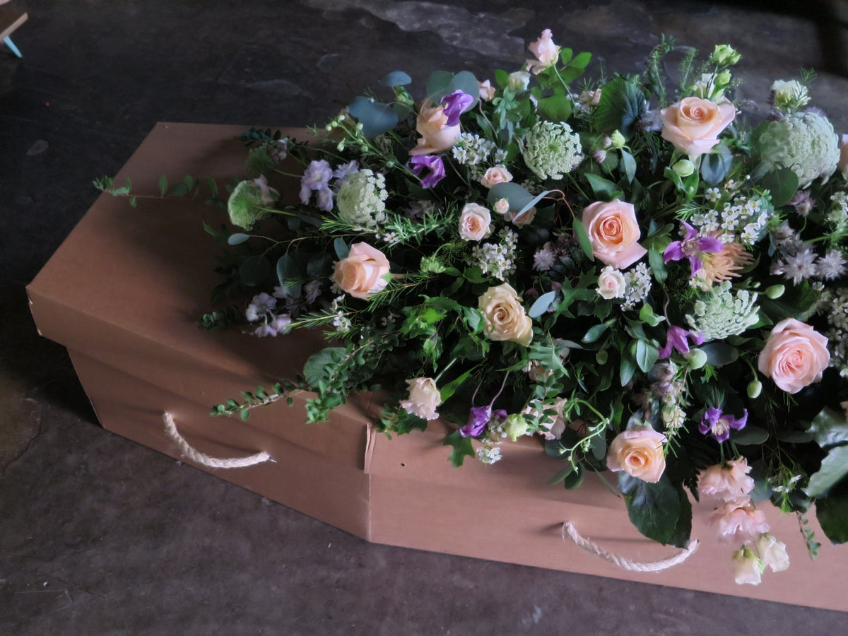 The 'From the Garden' Casket Spray by Wild About Flowers elegantly decorates a cardboard coffin with pink roses, purple blooms, and seasonal greenery, offering a heartfelt tribute on a dark floor.