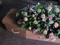 The 'From the Garden' Casket Spray by Wild About Flowers elegantly decorates a cardboard coffin with pink roses, purple blooms, and seasonal greenery, offering a heartfelt tribute on a dark floor.