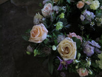 The 'From the Garden' Heart Design by Wild About Flowers showcases a lush wreath of flowers resting on a dark surface. It includes seasonal roses in shades of pale pink, peach, and cream, accented with greenery and small white blooms. The dim lighting creates soft shadows that enhance its enchanting allure.