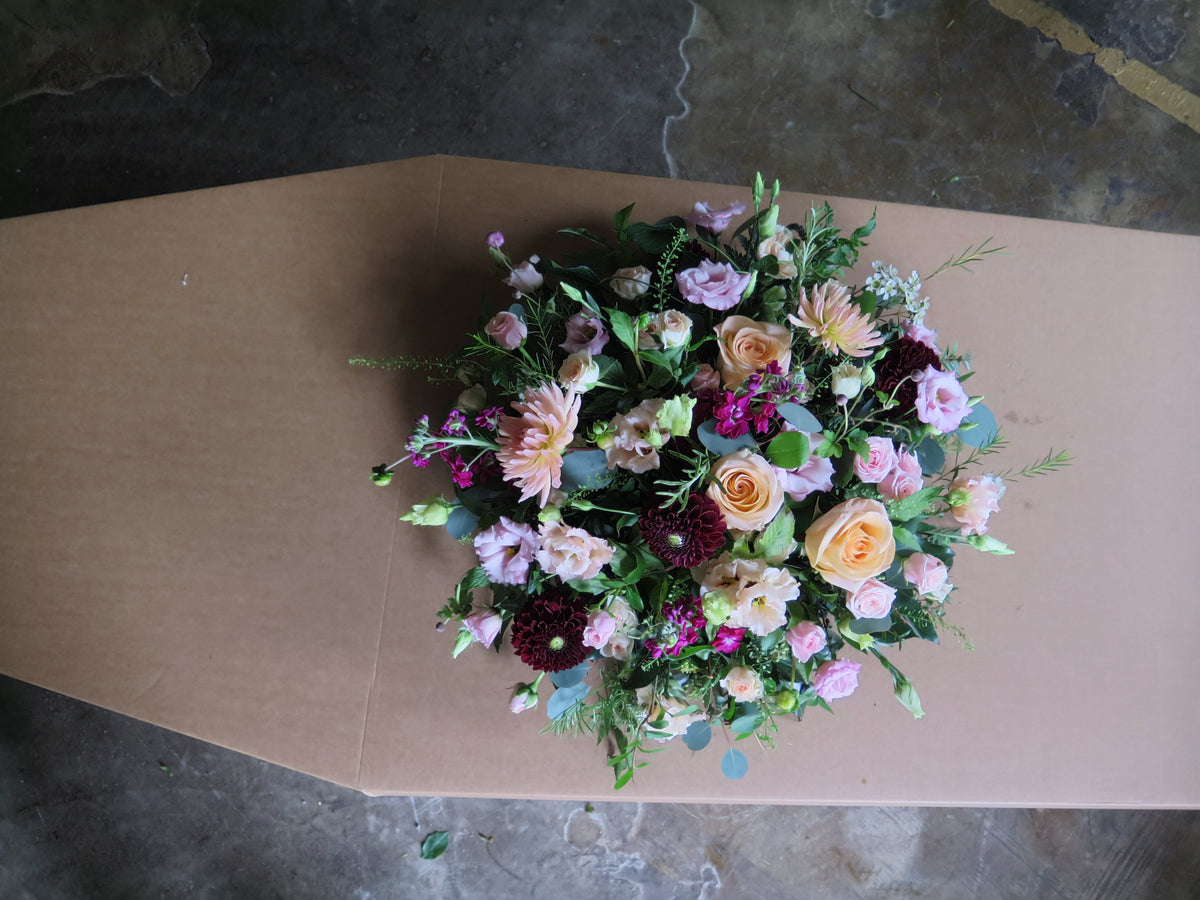 The 'From the Heart' Posy Design by Wild About Flowers is a sophisticated funeral piece showcasing a large, colorful floral arrangement atop a coffin. This elegant design features seasonal roses, dahlias, and fresh-cut foliage beautifully presented against a concrete floor backdrop.