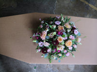 The 'From the Heart' Posy Design by Wild About Flowers is a sophisticated funeral piece showcasing a large, colorful floral arrangement atop a coffin. This elegant design features seasonal roses, dahlias, and fresh-cut foliage beautifully presented against a concrete floor backdrop.