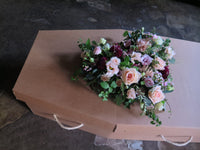 The 'From the Heart' Heart Design by Wild About Flowers, featuring a vibrant floral tribute with roses, scented herbs, and various blooms, adorns a cardboard coffin with rope handles as it rests on a textured stone floor.