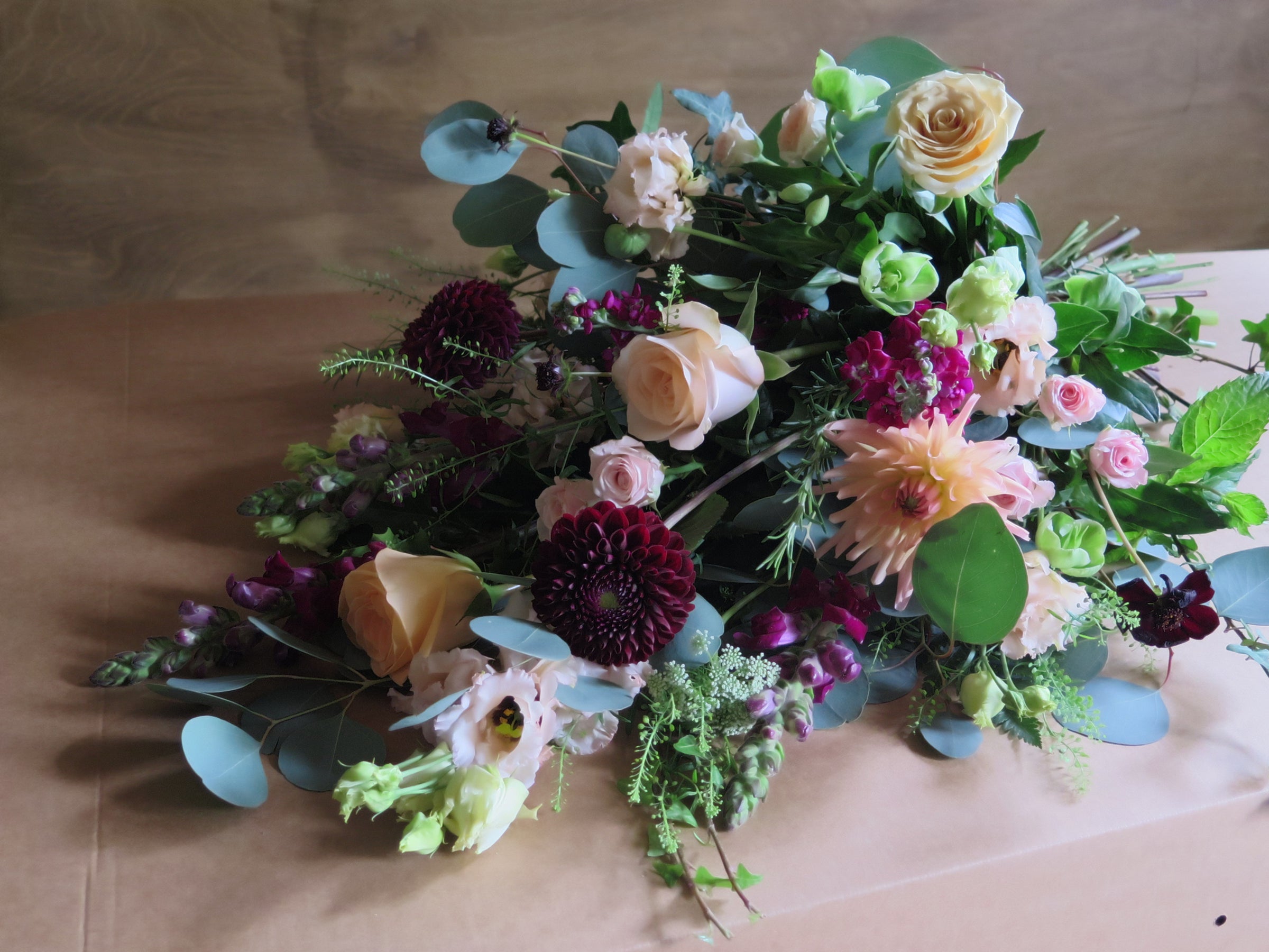 The 'From the Heart' Tied Sheaf by Wild About Flowers elegantly showcases pale pink roses, dark purple dahlias, and an assortment of greenery on a brown surface. This eco-friendly arrangement, with its blend of soft and rich tones, brings a sophisticated touch to any setting.