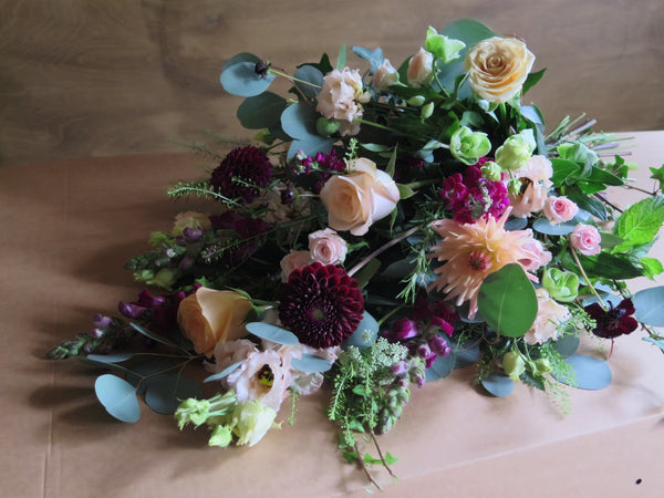 The 'From the Heart' Tied Sheaf by Wild About Flowers elegantly showcases pale pink roses, dark purple dahlias, and an assortment of greenery on a brown surface. This eco-friendly arrangement, with its blend of soft and rich tones, brings a sophisticated touch to any setting.