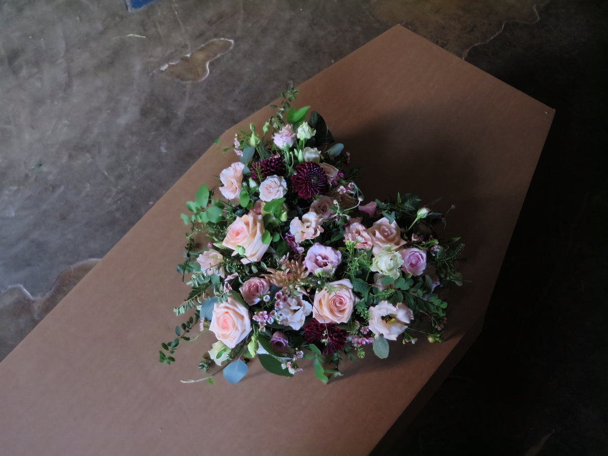 A coffin adorned with the 'From the Heart' Heart Design by Wild About Flowers, showcasing a floral tribute of pink roses, greenery, and dark maroon flowers arranged in a heart shape. The ambiance is dimly lit and somber, set against a concrete floor.