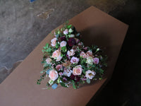 A coffin adorned with the 'From the Heart' Heart Design by Wild About Flowers, showcasing a floral tribute of pink roses, greenery, and dark maroon flowers arranged in a heart shape. The ambiance is dimly lit and somber, set against a concrete floor.