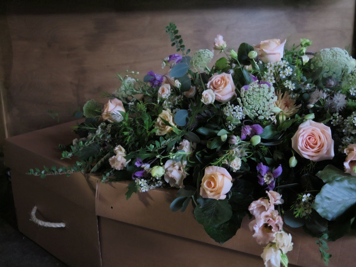 A wooden coffin embellished with the 'From the Garden' Casket Spray by Wild About Flowers, showcasing seasonal flowers like pink roses, white blooms, greenery, and purple accents. Soft lighting enhances a serene and respectful tribute atmosphere.