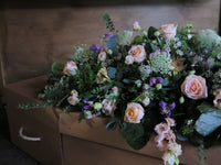 A wooden coffin embellished with the 'From the Garden' Casket Spray by Wild About Flowers, showcasing seasonal flowers like pink roses, white blooms, greenery, and purple accents. Soft lighting enhances a serene and respectful tribute atmosphere.