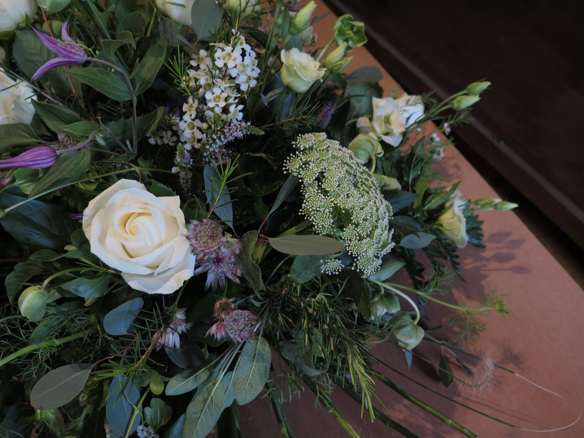 The 'From the Garden' Woodland Spray by Wild About Flowers exhibits an elegant arrangement of white roses and small white wildflowers interwoven with lush garden foliage, all artfully displayed on a wooden surface.