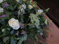 The 'From the Garden' Woodland Spray by Wild About Flowers exhibits an elegant arrangement of white roses and small white wildflowers interwoven with lush garden foliage, all artfully displayed on a wooden surface.
