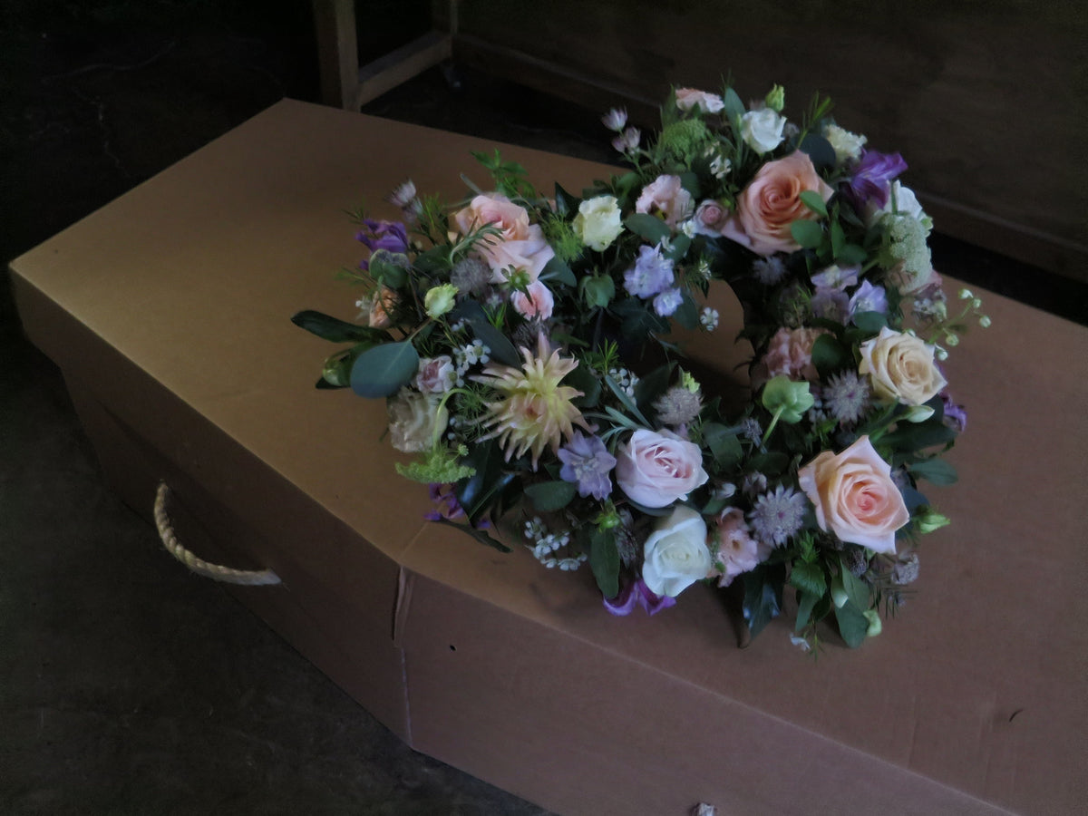 In a dimly lit room, a closed brown casket is adorned with the 'From the Garden' Heart Design wreath by Wild About Flowers, featuring seasonal pink roses, white flowers, and greenery.