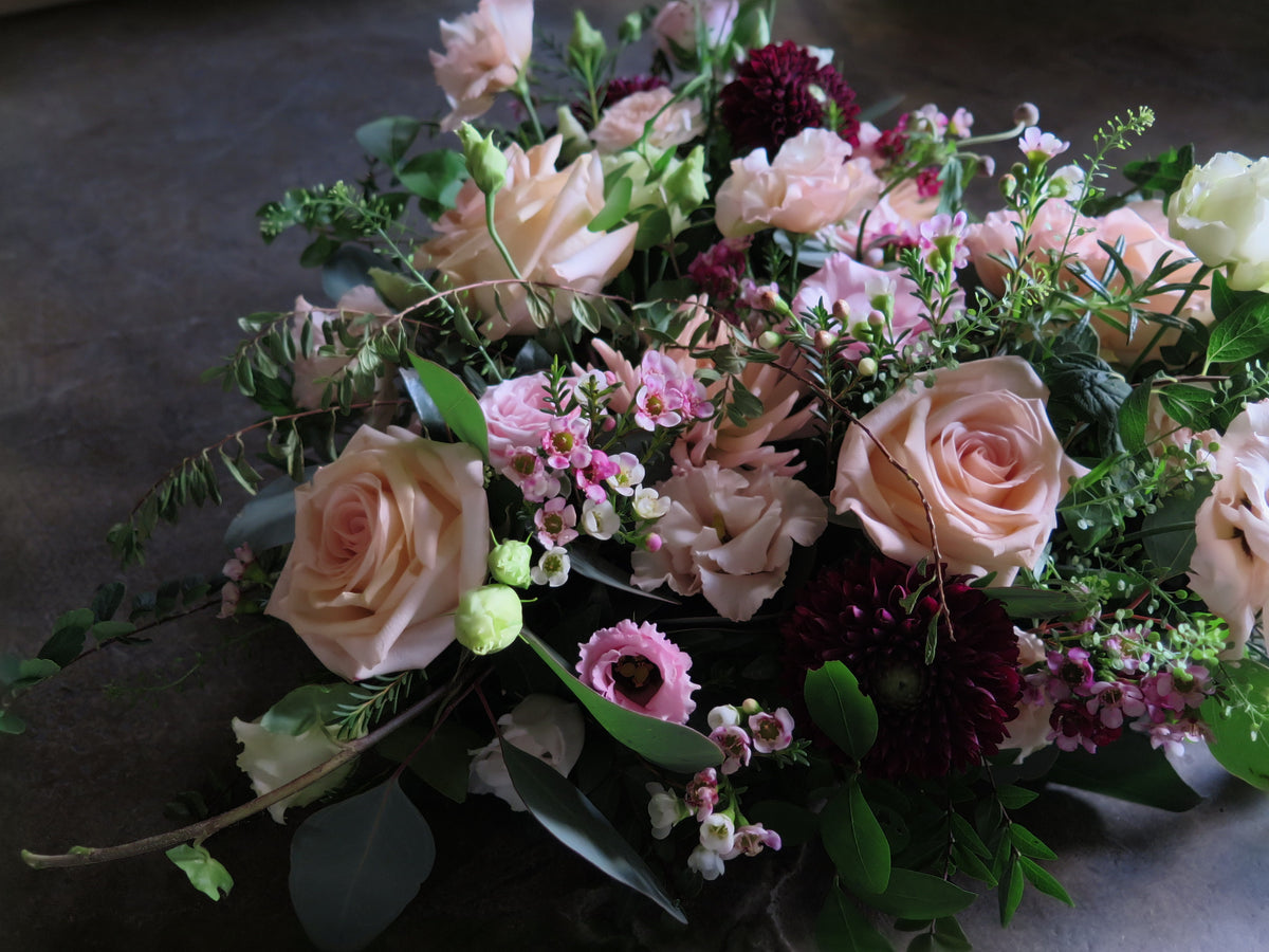 The 'From the Heart' Heart Design by Wild About Flowers is a lush floral arrangement showcasing pale pink roses, dark red chrysanthemums, and a mix of white and pink blooms, complemented by greenery and scented herbs, all set against a dark background.