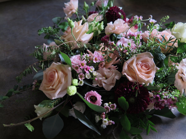 The 'From the Heart' Heart Design by Wild About Flowers is a lush floral arrangement showcasing pale pink roses, dark red chrysanthemums, and a mix of white and pink blooms, complemented by greenery and scented herbs, all set against a dark background.