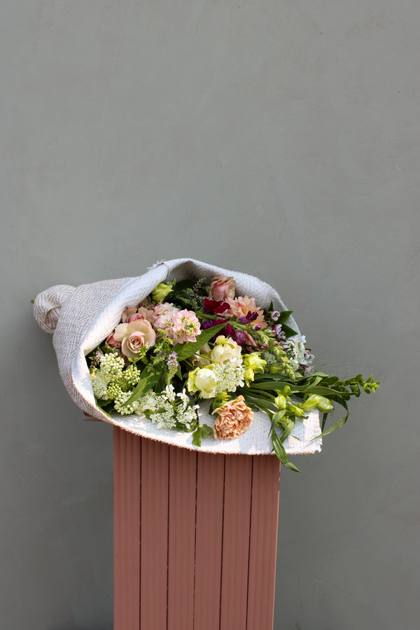 A floral wrap by Wild About Flowers, featuring seasonal blooms such as roses and carnations, is elegantly displayed on a wooden pedestal against a simple gray backdrop. The bouquet is wrapped in white paper and enhanced with lush greenery, creating a soft and timeless arrangement.