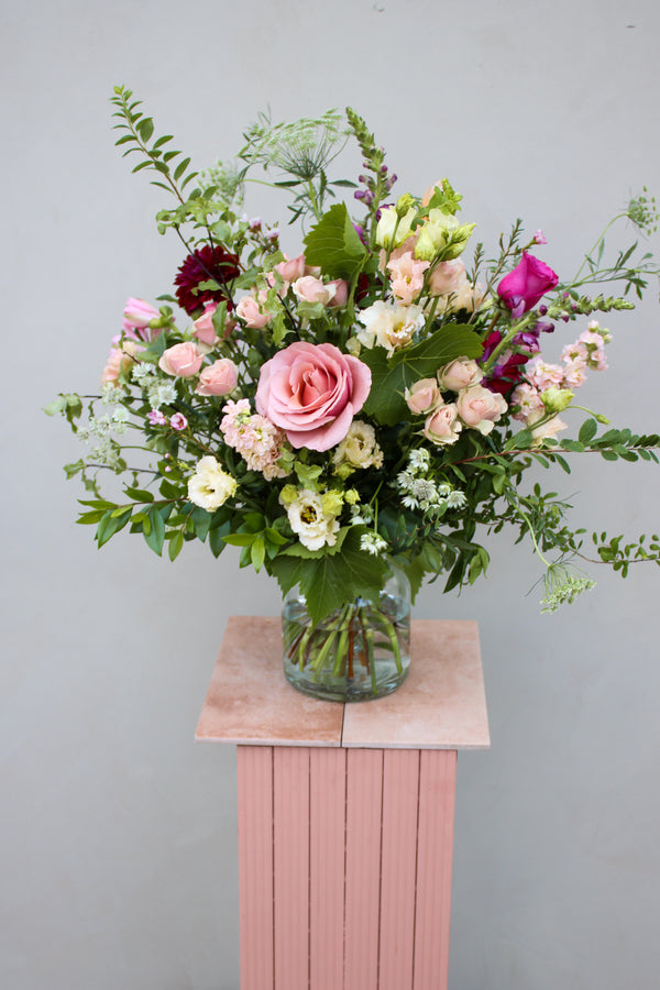 The Juicy Bunch by Wild About Flowers presents a seasonal flower bouquet in a glass vase, highlighting pink roses, white daisies, and lush greenery. This vivid arrangement is gracefully displayed on a pink pedestal against a neutral backdrop.