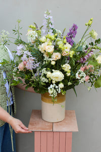 A person skillfully assembles a large floral bouquet in the Pretty Pot by Wild About Flowers, setting it on a pink pedestal. This breathtaking arrangement showcases roses, lisianthus, and greenery in shades of pink, white, and purple. A plain light gray wall serves as the backdrop, accentuating the elegance of this sustainable decor.