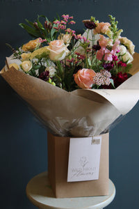 The "Wild About Flowers" bouquet, showcasing Event's floral design skills, features pink roses and wildflowers. It is wrapped in brown paper with a tag and placed in a cardboard box. Displayed against a dark blue wall, it hints at the Handtied Bouquet Flower Workshop in Bristol.
