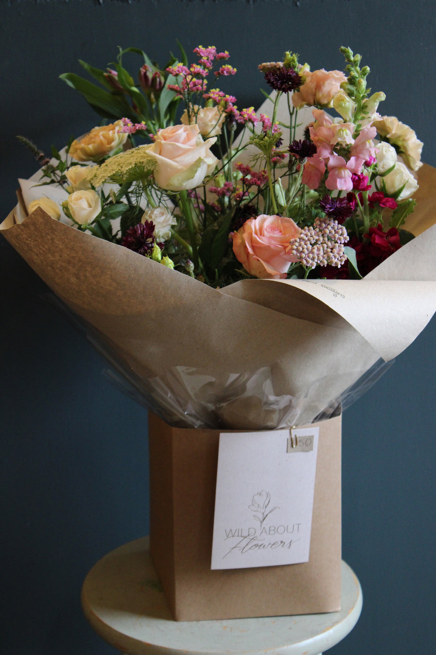 The "Wild About Flowers" bouquet, showcasing Event's floral design skills, features pink roses and wildflowers. It is wrapped in brown paper with a tag and placed in a cardboard box. Displayed against a dark blue wall, it hints at the Handtied Bouquet Flower Workshop in Bristol.