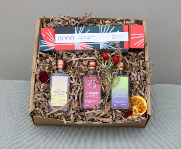 The Crackers about Gin Gift Box by Wild About Flowers is ideal for Christmas gifts. It includes three small bottles of Chew Valley Distillery gin and a box of chocolate buttons, all nestled in brown shredded paper. The festive presentation is completed with dried flowers, berries, and a slice of dried orange.