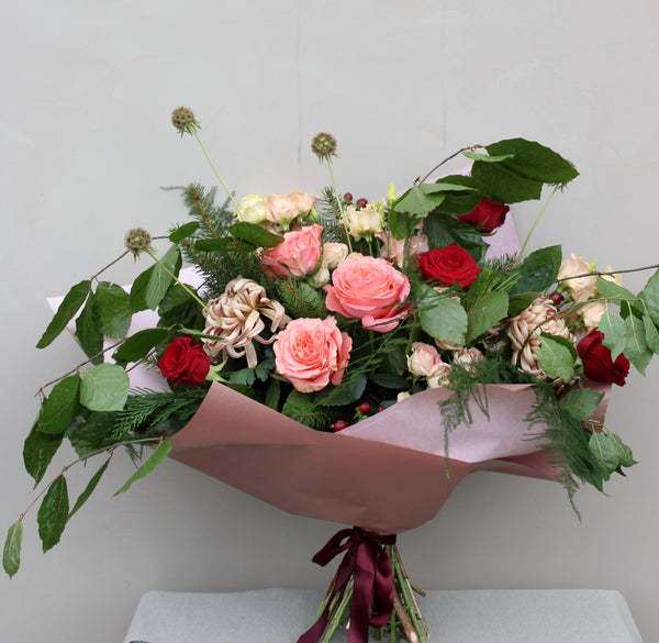 The Santa Baby Blooms by Wild About Flowers features a bouquet of pink and red roses, accented with greenery and wrapped in pink paper, tied with a dark red ribbon—a perfect touch for the festive season.