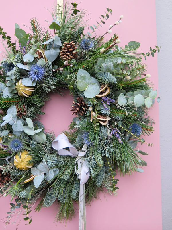 A striking Winter Wonderland Wreath by Wild About Flowers, crafted with evergreen branches, eucalyptus leaves, pinecones, thistles, and dried citrus slices adorned with a white ribbon, is showcased against a pink backdrop. This exquisite piece is ideal for your Christmas décor.