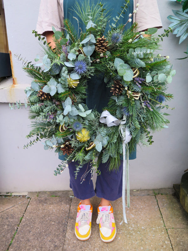 A person holding a handmade Winter Wonderland Wreath from Wild About Flowers, decorated with pinecones, eucalyptus leaves, and a silver ribbon. The individual is wearing a green apron and colorful sneakers while standing on a stone pavement near a light gray wall.