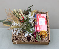 The Christmas at Home Gift Box by Wild About Flowers includes a beautiful bouquet of dried Christmas bunch flowers, a festive pink-wrapped chocolate bar from Coco Chocolatier, and a decorative dried orange slice, all thoughtfully arranged on shredded paper. The box is elegantly displayed on a textured green background.