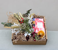 The "Christmas at Home Gift Box" by Wild About Flowers contains a festive dried Christmas bunch, a vibrant pink package marked "Coco," and a slice of dried orange. The box is filled with shredded paper and placed on a light gray surface, capturing the essence of holiday cheer in every detail.