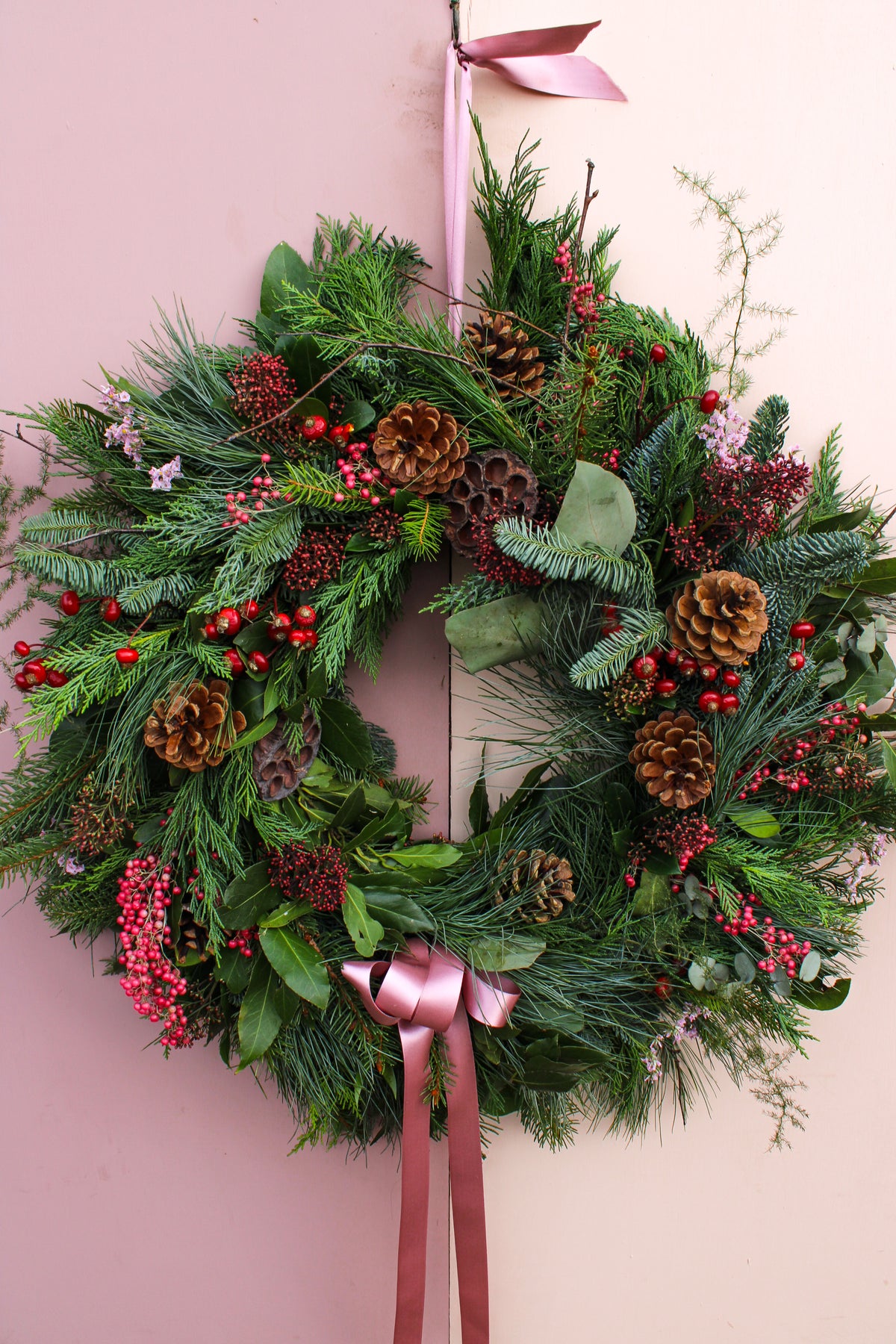 The Wild About Flowers Love Actually Winter Wreath, adorned with pinecones, red berries, green pine branches, and a pink ribbon bow, hangs beautifully on a light-colored door.